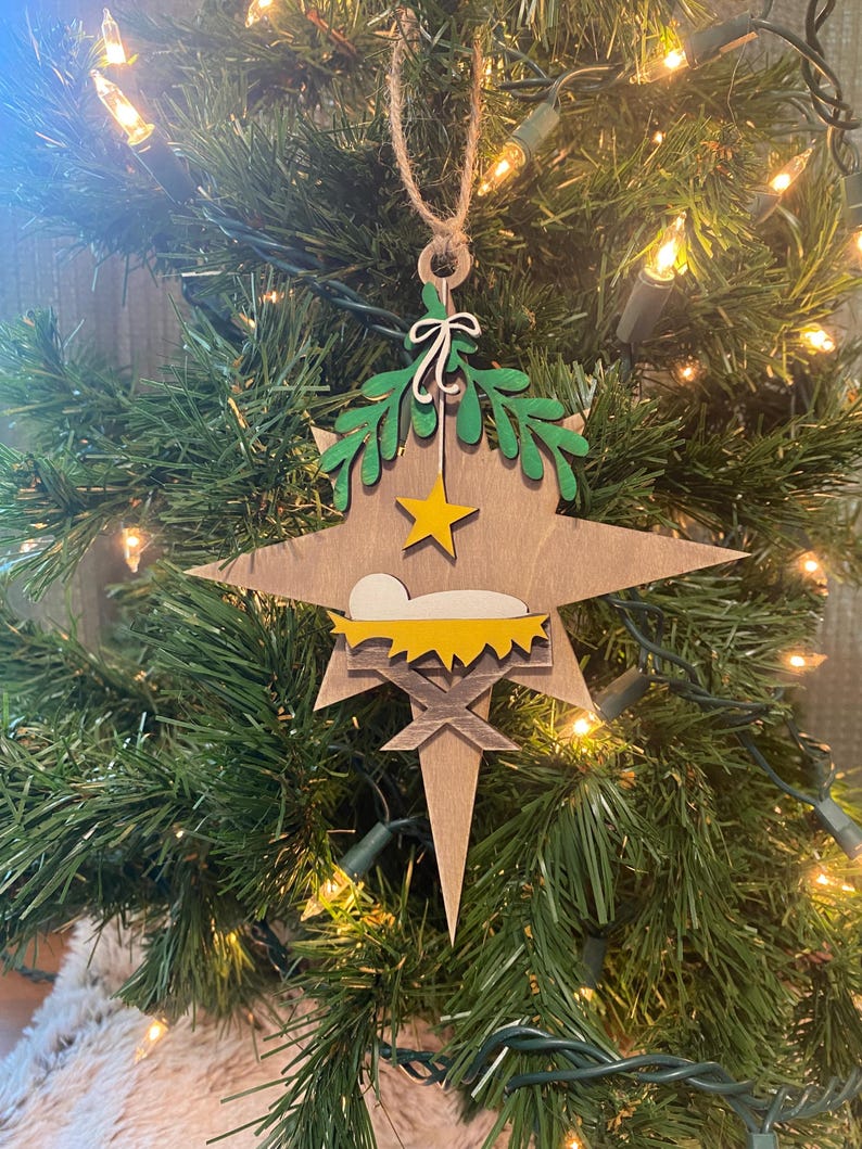 May include: A wooden Christmas ornament shaped like a star with a manger scene inside. The star is painted brown with a yellow star on top and green leaves around the edges. The manger is painted yellow with a white baby inside.