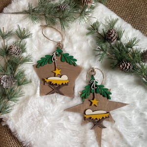 May include: Two wooden star-shaped Christmas ornaments with a manger scene and a star on each. The ornaments are painted in shades of brown, green, and yellow. The ornaments are hanging from a white fuzzy surface.