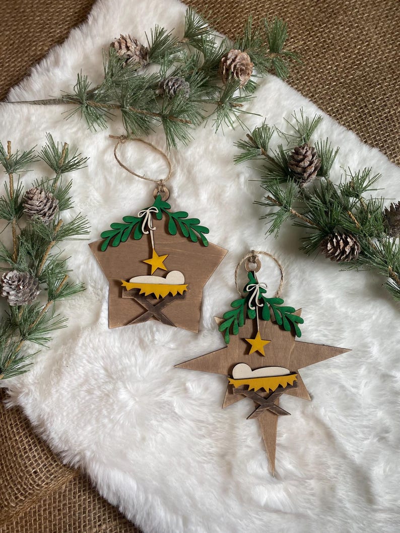 May include: Two wooden star-shaped Christmas ornaments with a manger scene and a star on each. The ornaments are painted in shades of brown, green, and yellow. The ornaments are hanging from a white fuzzy surface.