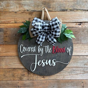 May include: A round wooden sign with the text "Covered by the Blood of Jesus" in white and red. It is decorated with a black and white checkered bow, green leaves, and a burlap backing. The sign hangs on a wooden wall.