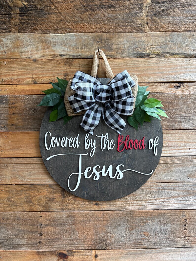May include: A round wooden sign with the text "Covered by the Blood of Jesus" in white and red. It is decorated with a black and white checkered bow, green leaves, and a burlap backing. The sign hangs on a wooden wall.