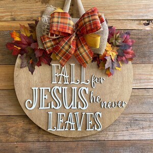May include: A round wooden door hanger with a fall theme. It features a burlap bow with a plaid ribbon, fall leaves, and the text "Fall for Jesus he never leaves".