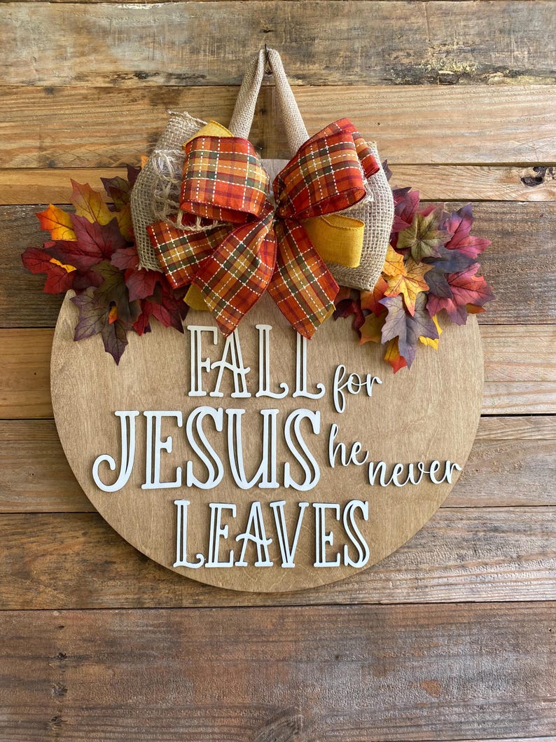 May include: A round wooden door hanger with a fall theme. It features a burlap bow with a plaid ribbon, fall leaves, and the text "Fall for Jesus he never leaves".