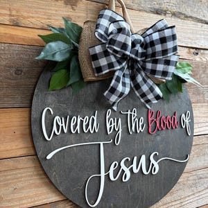 May include: A round wooden sign with the text "Covered by the Blood of Jesus" in white script and red accents. It is decorated with a burlap accent, a black and white checkered bow, and green leaves. The sign is hanging on a wooden background.