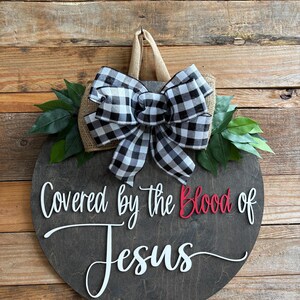 May include: A round wooden sign with the words "Covered by the Blood of Jesus" in white and red lettering. The sign is decorated with a black and white checkered bow, burlap, and green leaves. It has a jute hanger.