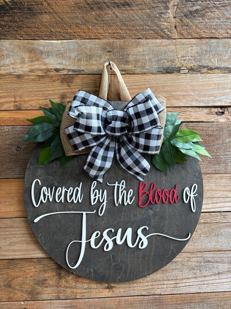 May include: A round wooden sign with the words "Covered by the Blood of Jesus" in white and red lettering. The sign is decorated with a black and white checkered bow, burlap, and green leaves. It has a jute hanger.