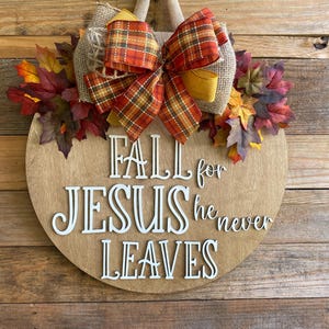 May include: A round wooden sign with the text "Fall for Jesus he never leaves" in white letters. The sign is decorated with a burlap bow with a plaid ribbon and fall leaves.
