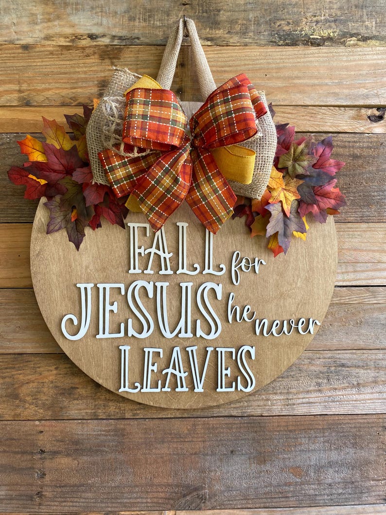May include: A round wooden sign with the text "Fall for Jesus he never leaves" in white letters. The sign is decorated with a burlap bow with a plaid ribbon and fall leaves.