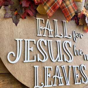 May include: A wooden round sign with the words "Fall for Jesus he never leaves" in white letters. The sign is decorated with a brown burlap bow, a plaid ribbon, and fall leaves.