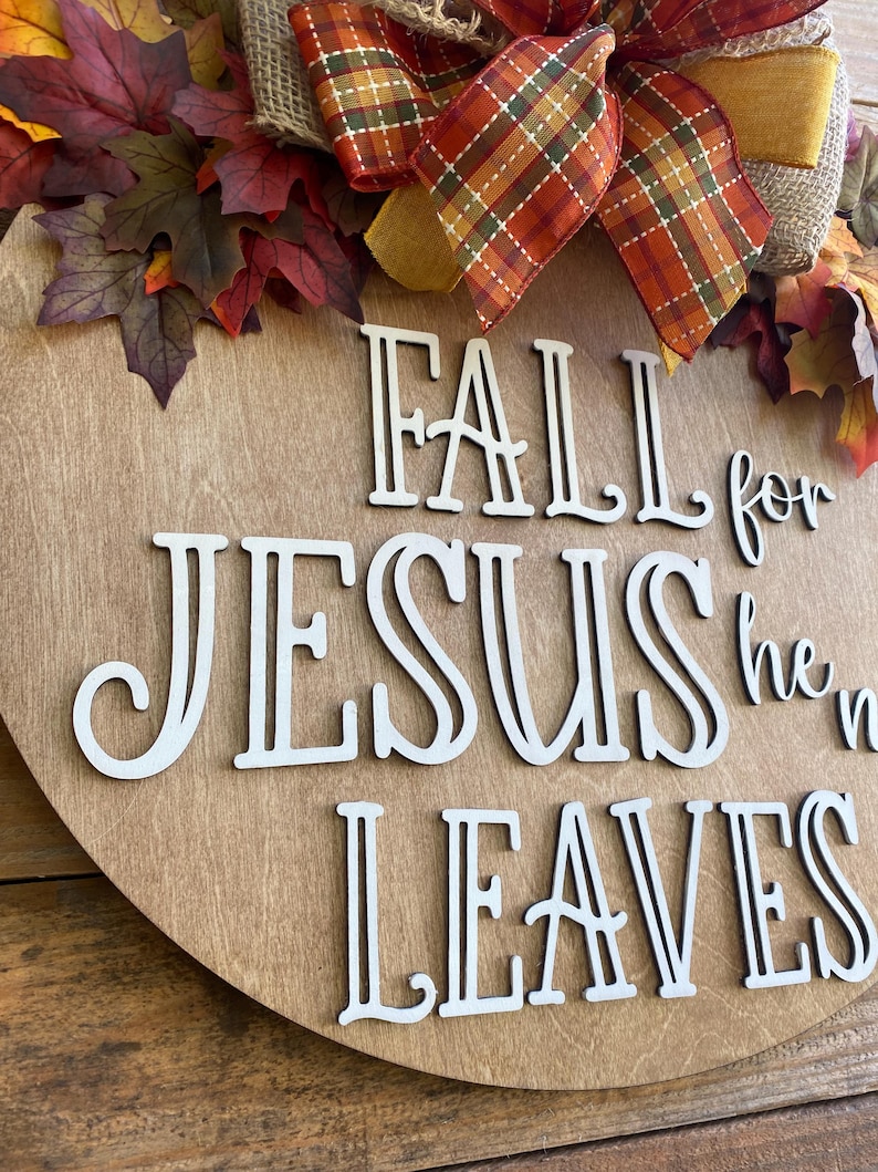 May include: A wooden round sign with the words "Fall for Jesus he never leaves" in white letters. The sign is decorated with a brown burlap bow, a plaid ribbon, and fall leaves.
