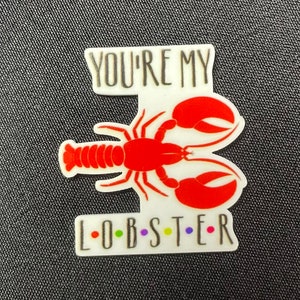 Pu&ograve; includere: Spilla a tema aragosta bianca e rossa con la scritta "YOU'RE MY L-O-B-S-T-E-R" in lettere nere e colorate. L'aragosta &egrave; rossa con dettagli bianchi. La spilla &egrave; su una superficie strutturata grigio scuro.