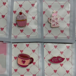 Może przedstawiać: Kolekcja emaliowanych przypinek walentynkowych z różnymi wzorami. Zawiera babeczkę z wiśnią, czajniczek z serduszkami, słoik wypełniony sercami, butelkę wina, kubek z sercem i usta z napisem "Kiss Me".