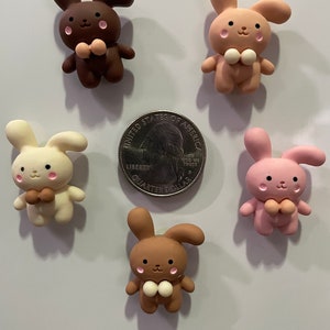 Könnte beinhalten: Sechs hasenförmige Anhänger in verschiedenen Farben: Dunkelbraun, Hellbraun, Creme und Rosa. Jeder Anhänger hat eine Fliege und rosige Wangen. Eine Münze dient als Größenvergleich. Diese Anhänger sind wahrscheinlich für Bastelarbeiten oder Schmuckherstellung gedacht.