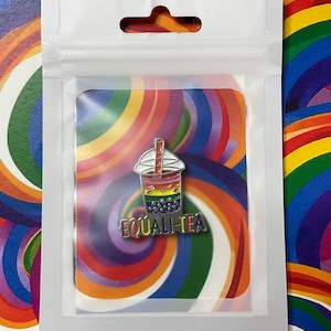 Può includere: Una spilla smaltata colorata in un sacchetto di plastica trasparente. La spilla presenta un design di tè alle bolle arcobaleno con la scritta "EQUALITEA". Lo sfondo è un vortice di colori arcobaleno.
