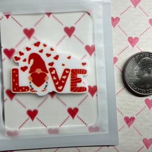 Könnte beinhalten: Ein Valentinstag-Gnom-Aufkleber mit dem Wort "LOVE" in Rot und Weiß. Der Gnom hält ein rotes Herz, und das Design enthält rote Herzen. Der Aufkleber befindet sich in einer transparenten Plastikhülle.