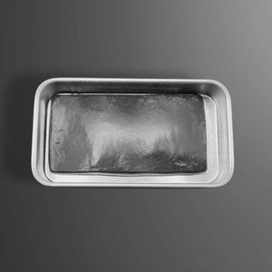 May include: A rectangular, dark gray object sits inside a silver-colored, rectangular tray. The object has a textured surface, and the tray has rounded corners. The background is a gradient of gray.