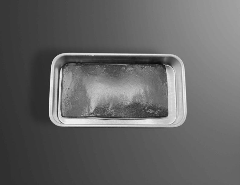 May include: A rectangular, dark gray object sits inside a silver-colored, rectangular tray. The object has a textured surface, and the tray has rounded corners. The background is a gradient of gray.