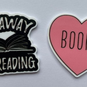 Może przedstawiać: Dwie naklejki: jedna czarno-biała z napisem „GO AWAY I'M READING” nad otwartą książką i różowa naklejka w kształcie serca ze słowem „BOOKS” w kolorze czarnym.