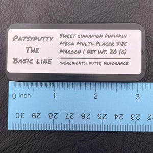 Op de afbeelding: Een rechthoekige zwarte blikken doos met het label Patsyputty The Basic Line Sweet Cinnamon Pumpkin Mega Multi-Placer Size Maroon Net Wt. 20 (g) Ingredients: Putty, Fragrance. Een blauwe liniaal toont een lengte van ongeveer 8,9 cm.