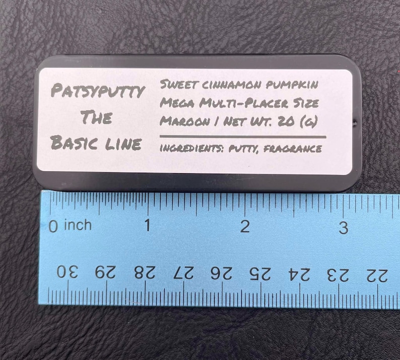 Op de afbeelding: Een rechthoekige zwarte blikken doos met het label Patsyputty The Basic Line Sweet Cinnamon Pumpkin Mega Multi-Placer Size Maroon Net Wt. 20 (g) Ingredients: Putty, Fragrance. Een blauwe liniaal toont een lengte van ongeveer 8,9 cm.