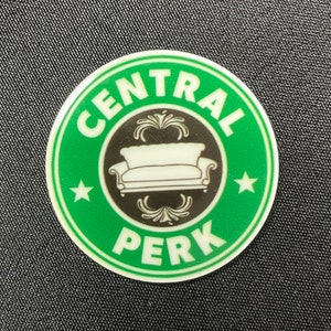 Pu&ograve; includere: Un adesivo rotondo verde e bianco con il logo Central Perk e un centro nero. Il logo presenta un divano bianco e le parole "CENTRAL PERK" e "PERK" in bianco. Due stelle bianche ai lati del divano.