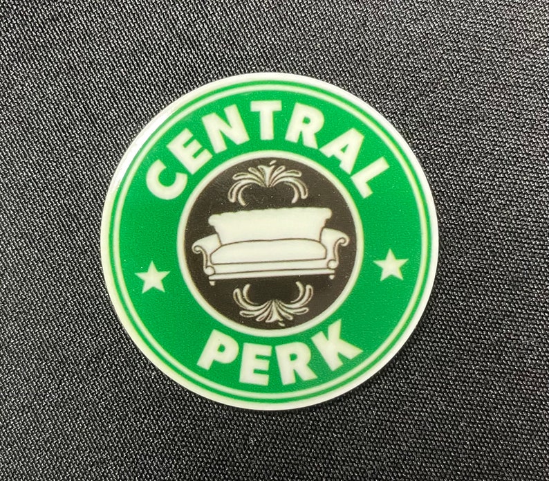 Pu&ograve; includere: Un adesivo rotondo verde e bianco con il logo Central Perk e un centro nero. Il logo presenta un divano bianco e le parole "CENTRAL PERK" e "PERK" in bianco. Due stelle bianche ai lati del divano.