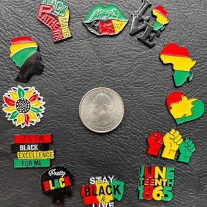 May include: A collection of colorful enamel pins with Black pride themes. Designs include a fist, lips, an African continent outline, and text like "Black Fathers Matter," "It's the Black Excellence for Me," and "June Teenth 1865." A US quarter is included for size reference.
