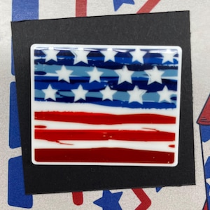 Puede incluir: Un pin rectangular con el diseño de la bandera estadounidense. El pin tiene un borde blanco, con un campo azul de estrellas blancas y rayas rojas y blancas. El pin está sobre un fondo negro.