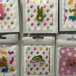 May include: A collection of enamel pins featuring bunny designs in various colors, including pink, green, and yellow. Each pin is displayed on a card with a paw print or polka dot pattern, packaged in a clear plastic sleeve.