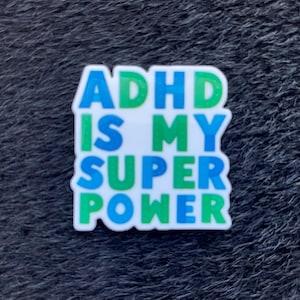 Könnte beinhalten: Ein weißer Anhänger mit dem Text "ADHD IS MY SUPER POWER" in blauen und grünen Buchstaben. Der Anhänger befindet sich auf einer dunklen, strukturierten Oberfläche.