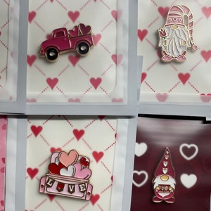 Könnte beinhalten: Eine Sammlung von rosa Emaille-Anstecknadeln mit goldenen Akzenten, die sich dem Valentinstag widmen. Designs umfassen ein Haus, einen Truck, Zwerge, ein Auto und eine Herzkiste. Jede Nadel wird auf einem Hintergrund mit Herzmuster präsentiert.