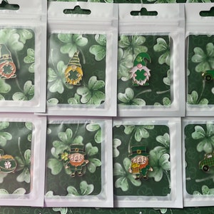 Może przedstawiać: Kolekcja emaliowanych przypinek na Dzień Świętego Patryka w przezroczystych plastikowych opakowaniach. Przypinki przedstawiają gnomy, skrzaty i zieloną ciężarówkę, wszystkie w zielonej, złotej i czerwonej kolorystyce. Tło to wzór zielonej koniczyny.