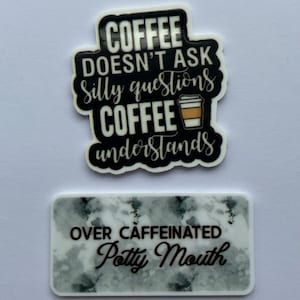 Könnte beinhalten: Zwei Aufkleber, einer oval und einer rechteckig. Der ovale Aufkleber trägt den Text "COFFEE DOESN'T ASK Silly questions COFFEE understands" mit einer Kaffeetassen-Grafik. Der rechteckige Aufkleber lautet "OVER CAFFEINATED Petty Mouth".