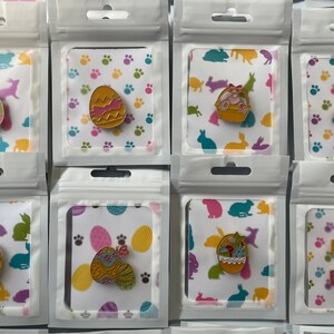 May include: A collection of Easter-themed enamel pins in clear plastic packaging. The pins feature colorful egg designs, Easter baskets, and a wreath with "Happy Easter" text. The background cards have bunny and paw print patterns.