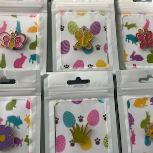 May include: A collection of enamel pins in clear plastic packaging. The pins feature various designs, including butterflies, flowers, grass, and a sun. The background of each package has a pattern of Easter eggs, paw prints, and bunnies.