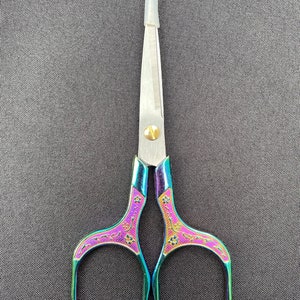 Könnte beinhalten: Kleine, dekorative Schere mit schillerndem Regenbogen-Finish. Die Griffe sind mit floralen Mustern in Lila und Rosa verziert. Die Klingen sind silberfarben und die Schere hat eine goldfarbene Schraube.