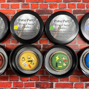 May include: Eight round tins of "PatsyPutty Wizard Sugar Cookies" are displayed against a brick wall. Each tin contains a decorated sugar cookie. The cookies are red, yellow, green, and blue, with wizard-themed decorations.