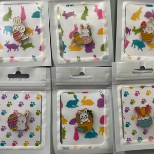 Może przedstawiać: Sześć emaliowanych przypinek z motywami wielkanocnych króliczków. Każda przypinka jest zapakowana w przezroczystą plastikową torebkę z ozdobną kartą. Przypinki przedstawiają króliki z pisankami i w koszykach, w różnych kolorach.