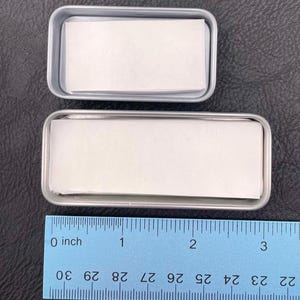 Op de afbeelding: Twee rechthoekige zilverkleurige metalen blikjes met stapels wit papier. Het kleinere bovenste blikje is ongeveer 6,4 cm lang, terwijl het grotere onderste blikje ongeveer 8,9 cm lang is, zoals aangegeven op de blauwe liniaal.