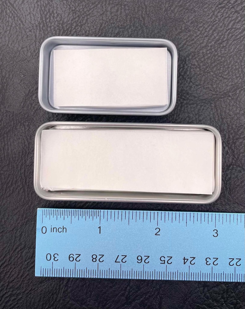 Op de afbeelding: Twee rechthoekige zilverkleurige metalen blikjes met stapels wit papier. Het kleinere bovenste blikje is ongeveer 6,4 cm lang, terwijl het grotere onderste blikje ongeveer 8,9 cm lang is, zoals aangegeven op de blauwe liniaal.