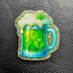 Könnte beinhalten: Ein dekorativer Artikel zum Thema St. Patrick's Day mit einem grünen Bierkrug mit weißem Schaum und Kleeblättern. Der Krug hat einen blauen Griff und Boden mit einem glitzernden grünen Rand. Das Design ist lebendig und auffällig.