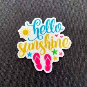 Puede incluir: Un charm blanco con las palabras "hello sunshine" en escritura azul y amarilla. El charm presenta un sol amarillo, chanclas rosas y estrellas y corazones coloridos, perfecto para el verano.