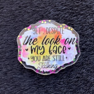 Könnte beinhalten: Ein klarer, unregelmäßig geformter Acryl-Pin mit dem Text "YET. DESPITE the look on my face you are still talking." Der Hintergrund zeigt Aquarellspritzer in Gelb, Orange und Blau. Der Pin ist mit rosa und gelbem Glitzer umrandet.