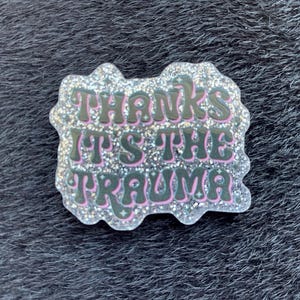 Könnte beinhalten: Ein glitzernder Anstecker mit dem Schriftzug "Thanks It's The Trauma" in einer Retro-Schriftart. Der Text ist dunkelgrün mit rosa Umrandung, vor silbernem Glitzerhintergrund.