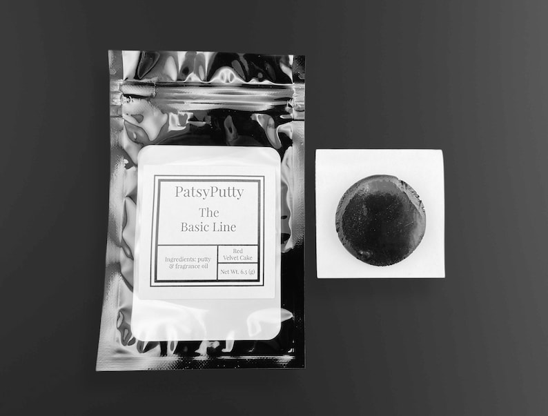 Puede incluir: Imagen en blanco y negro que muestra un paquete de PatsyPutty de The Basic Line, con informaci&oacute;n sobre ingredientes y aroma. Al lado, un peque&ntilde;o objeto oscuro y redondo descansa sobre un cuadrado blanco. El producto se identifica como Red Velvet Cake.