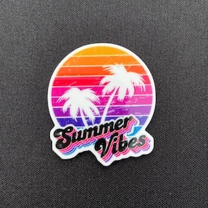Könnte beinhalten: Ein runder, farbenfroher Aufkleber mit einem Sonnenuntergangsdesign und zwei weißen Palmen. Der Text "Summer Vibes" ist in einer Retro-Schriftart mit blauem und rosa Umriss gehalten. Der Aufkleber befindet sich auf schwarzem Hintergrund.