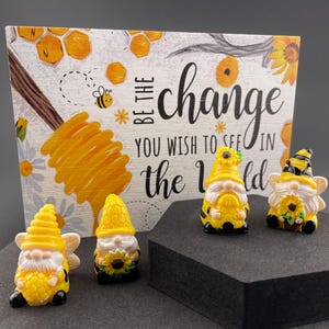 Könnte beinhalten: Vier gelb-schwarze Zwergenfiguren mit Bienen- und Sonnenblumen-Details werden vor einem Holzschild ausgestellt. Das Schild trägt den Text "BE THE CHANGE YOU WISH TO SEE IN THE WORLD" mit Bienen- und Waben-Grafiken.