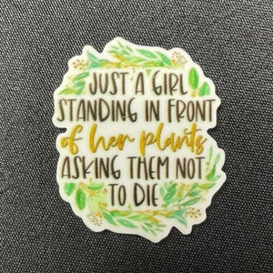 Könnte beinhalten: Weißer Aufkleber mit dem Text "JUST A GIRL STANDING IN FRONT of her plants ASKING THEM NOT TO DIE." Der Text ist in Schwarz und Gold gehalten, mit grünen Blättern. Der Aufkleber befindet sich auf einer dunkelgrauen, strukturierten Oberfläche.