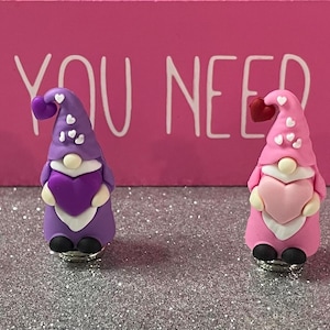 Könnte beinhalten: Vier Valentinstags-Wichtel in Rosa, Lila und Weiß, jeder hält ein rotes Herz. Die Wichtel haben spitze Hüte und stehen vor einem rosa Hintergrund mit dem Text "ALL YOU NEED IS LOVE".