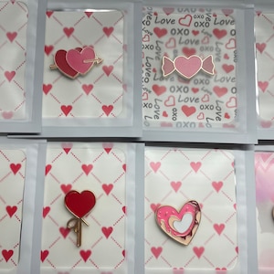 Könnte beinhalten: Eine Sammlung von Emaille-Anstecknadeln zum Valentinstag. Die Anstecknadeln zeigen Herzmotive in verschiedenen Farben, darunter Rot und Rosa, mit goldfarbenen Metallakzenten. Einige Anstecknadeln haben Text, andere Pfeile oder Vogelmotive.